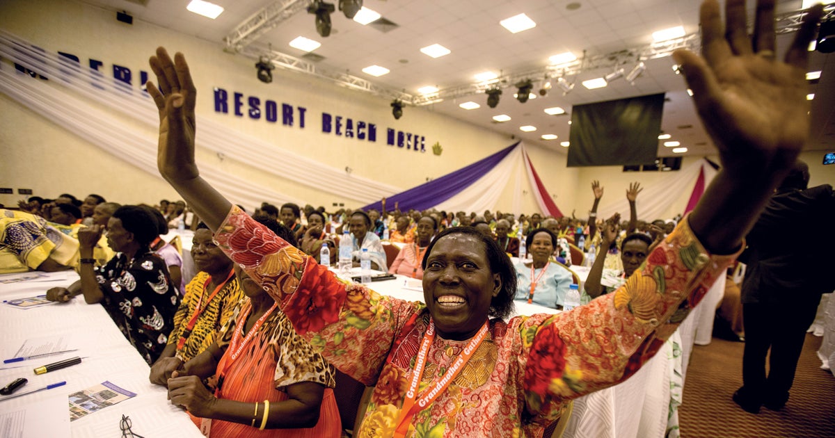 Smiling Ugandan woman with her arms outstretched with women seated in rows of tables behind her