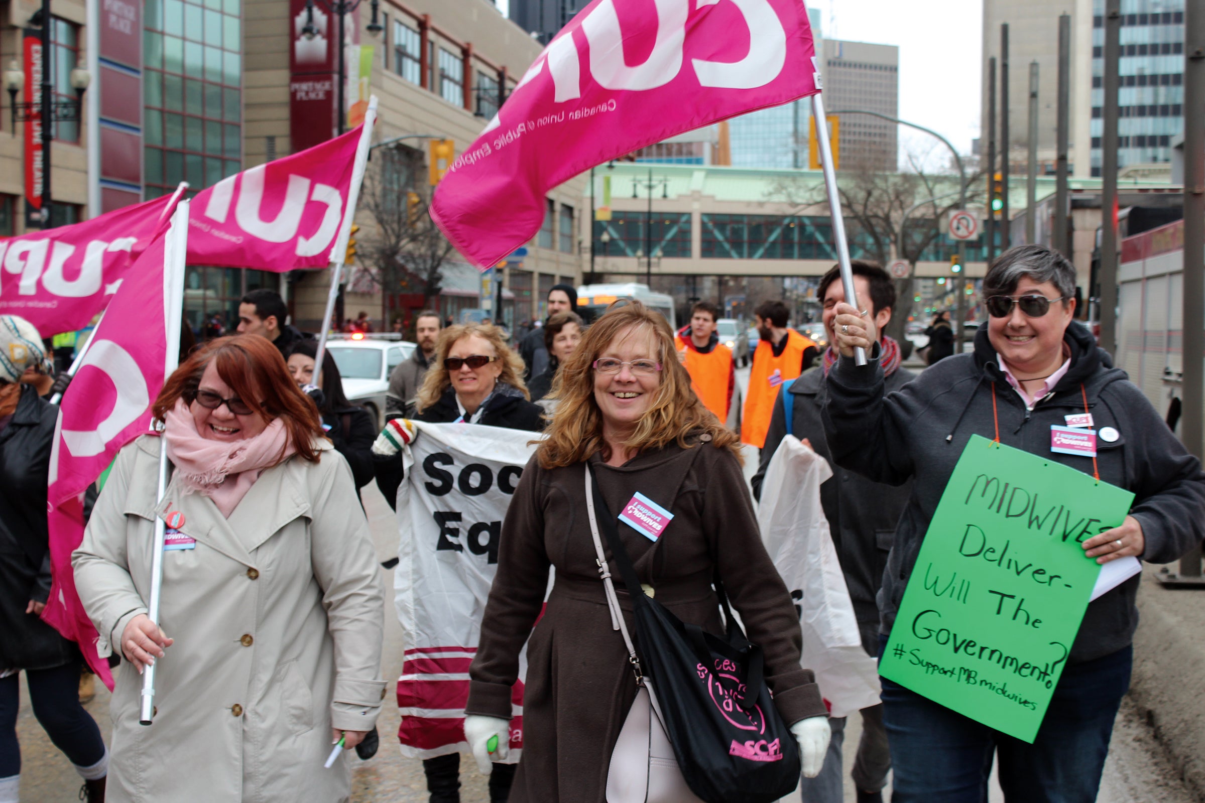 Women march holding CUPE flags and a protest sign that says &quot;Midwives deliver: will the government?&quot;