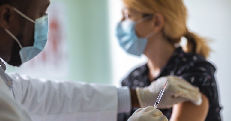 A masked healthcare worker swabs the arm of a patient to prepare for a vaccination