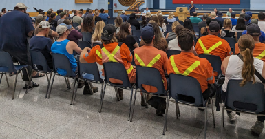 Workers sitting on chairs in an auditorium while a person speaks at a podium