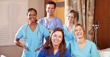 Young woman in blue scrubs standing beside bed holding a cup of orange liquid to the mouth of an elderly woman 