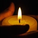 Two hands holding a small lit candle with darkness in the background