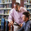 Two multi-ethnic men in the library looking at a computer screen and conversing. A mature man in his 40s is sitting at the desktop PC and a senior African-American man in his 60s is standing beside him.