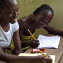 UN Mission Assists Women in Haiti Two Black women at a wooden desk writing with pencils on pads of paper