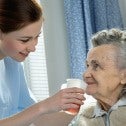 Young woman in blue scrubs standing beside bed holding a cup of orange liquid to the mouth of an elderly woman 