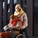 Man in wheel chair with woman standing behind him both wearing masks and winter clothes