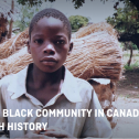 A young Black boy looks directly out of the frame, there are trees and a bundle of straw behind him