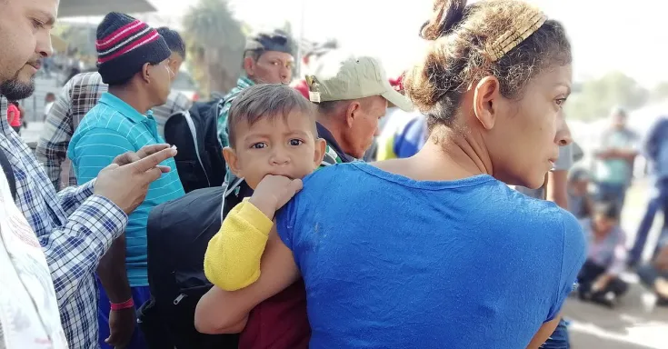 Picture shows a crowd of people, a woman in the foreground looking to the side, a child on her hip looks straight out at the viewer