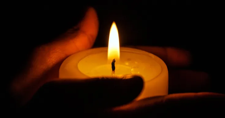 Two hands holding a small lit candle with darkness in the background