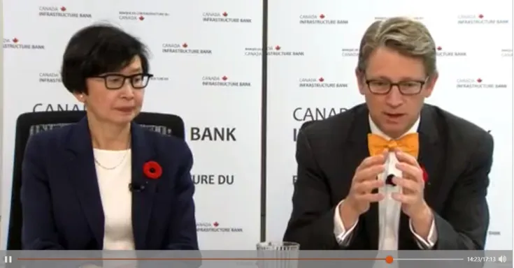 A woman in a navy blue suit jacket, a man in dark grey suit jacket and orange bowtie.