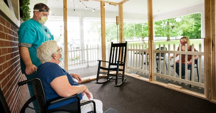 Long-term care worker in a mask and face shield stands next to an elder mask-wearing woman sitting in a wheelchair, speaking to a masked woman sitting outside a screened-in porch.