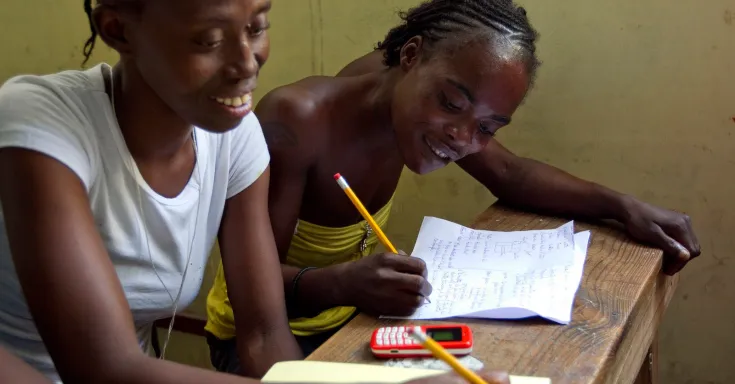 Two Black women at a wooden desk writing with pencils on pads of paper