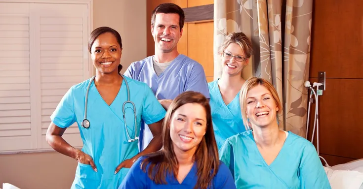 Young woman in blue scrubs standing beside bed holding a cup of orange liquid to the mouth of an elderly woman 