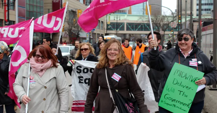 Women march holding CUPE flags and a protest sign that says "Midwives deliver: will the government?"