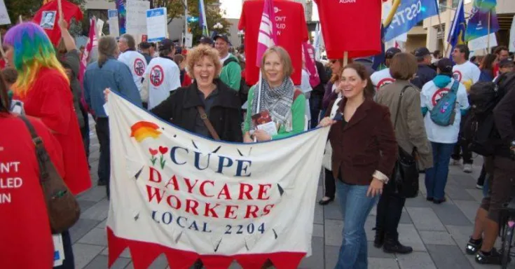 Three smiling white women holding a banner in a large crowd