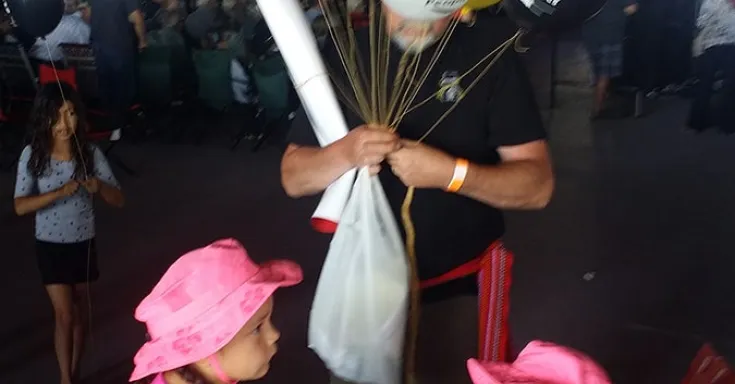 Man holding red, yellow, red and black balloons with two little girls in pink dresses and hats.