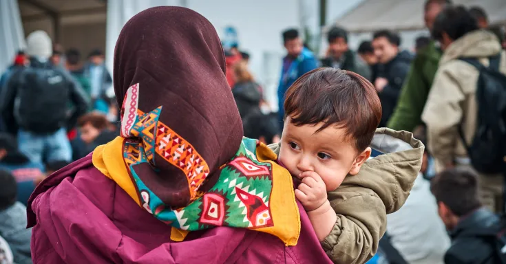 Woman with a colourful scarf holding a baby who looks over her shoulder at the camera.