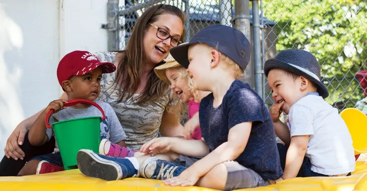 Four pre-school aged children laughing with a woman in glasses