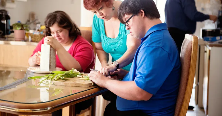 A woman standing at a table, leaning over to show a young man how to cut vegetables, a second woman at the table uses a cheese grater
