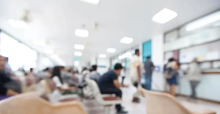 Blurry photo of people sitting and standing in a bright hospital waiting room
