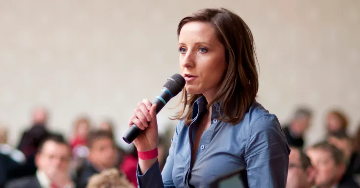 30ish white woman in a blue shirt holding a microphone and speaking