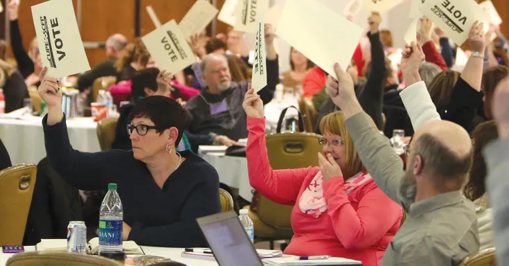 Roomful of people seated at tables holding up voter cards